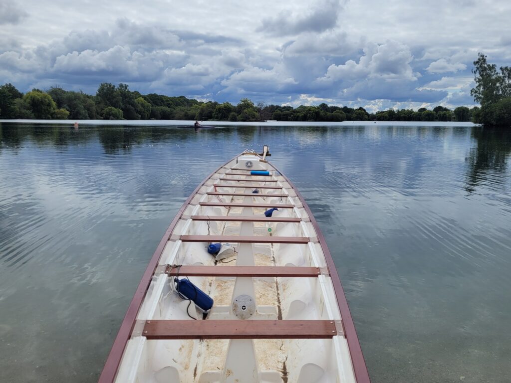 The dragonboat is empty sitting in the water after everyone has gotten out. The lake water is still and looks like glass. There are trees around the lake endge in the background.