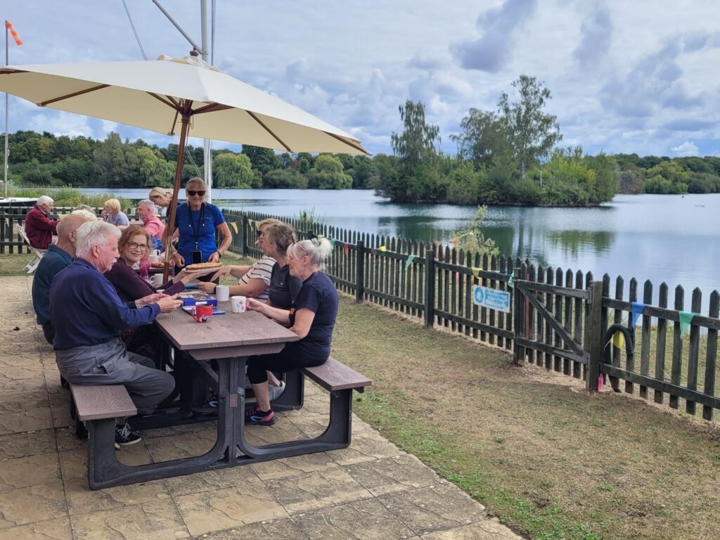 The OPfL team are sitting on picnic benches under a large parasol enjoying tea and home-made cake. The lake is on the right-hand side, the water is very still and there are trees along the lake short in the distance.