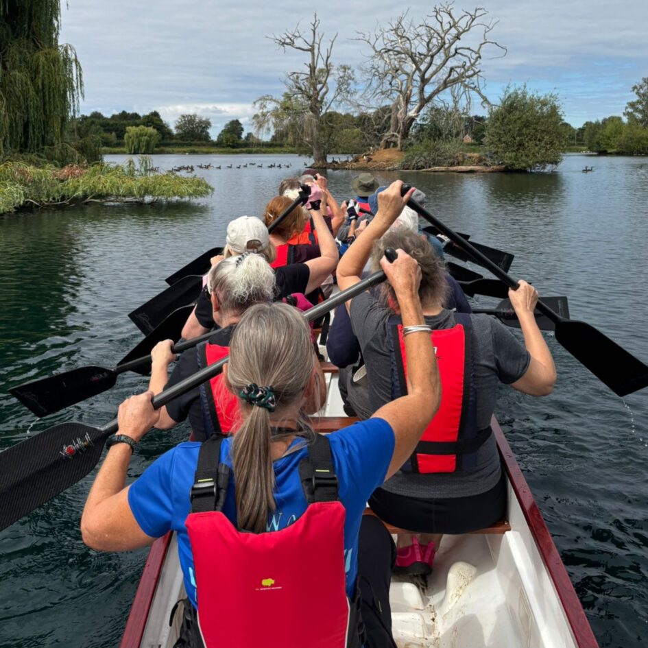 View of the boat from the back. Paddlers are ready with their paddles in the air. The lake is to the front, with small islands and geese.