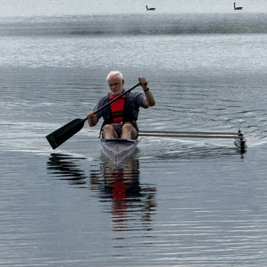 Man paddling in 'O1' outrigger boat. Paddle is in the water to the left, the outrigger float is to the right. The background is the lake with two geese in the distance.