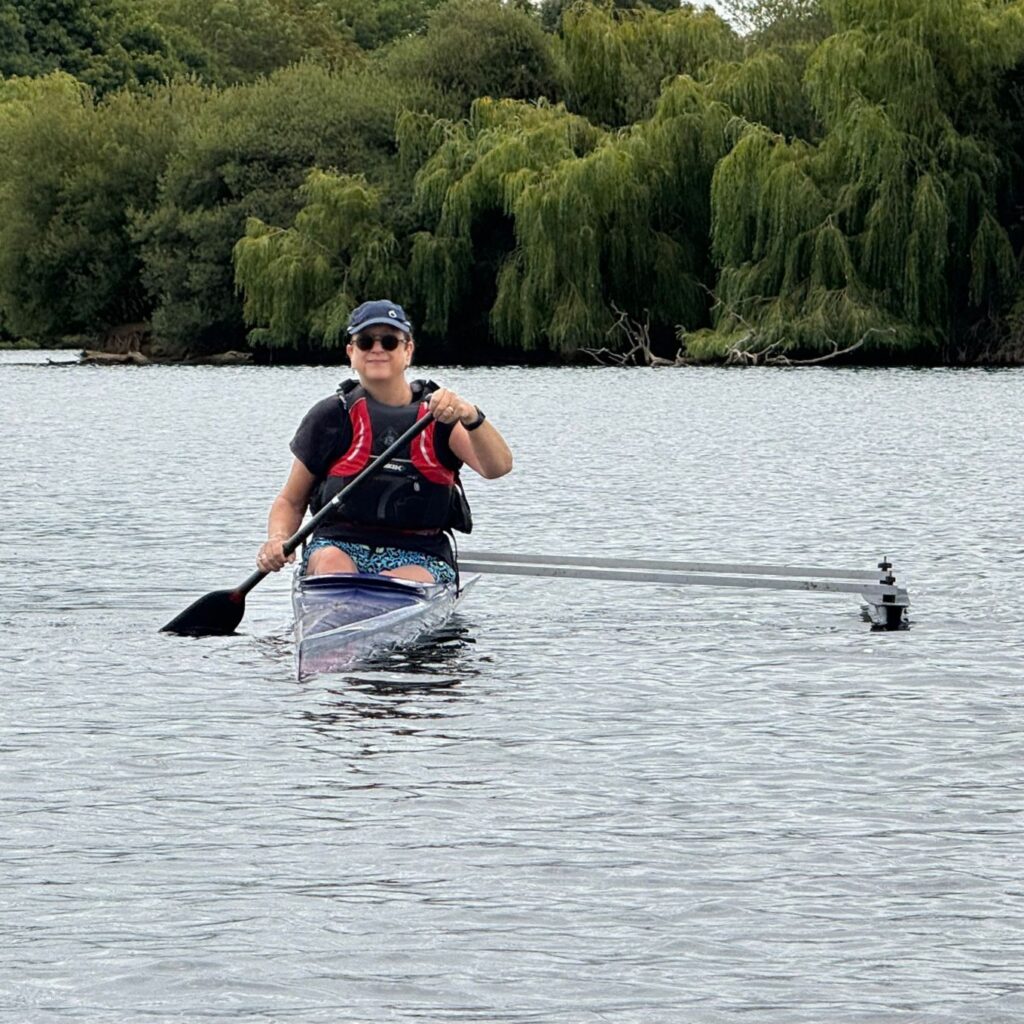 Female paddler in 'O1' outrigger boat, smiling at the camera. Paddle is in the water to the left, the outrigger float is to the right. Willow trees line the lake in the background.