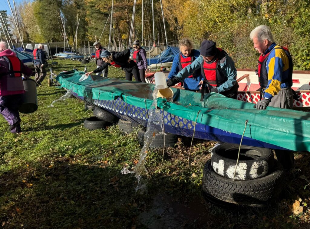 The dragonboat has a cover on it which has collected a lot of water. Paddlers are around the boat using scoops to bail out the water.