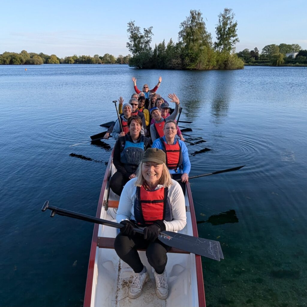 Photo taken looking from the front to the back of the boat. People have their paddles out to the side and are waving at the camera. The sky is blue, and so is the lake. Everyone looks happy.