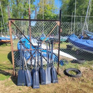 Dragonboat paddles stacked against the boatyard gate. There are small boats in the background, trees and blue sky.