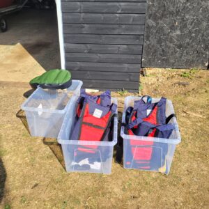 Plastic boxes containing buoyancy aids are in plastic boxes on the ground waiting for paddles to collect them.