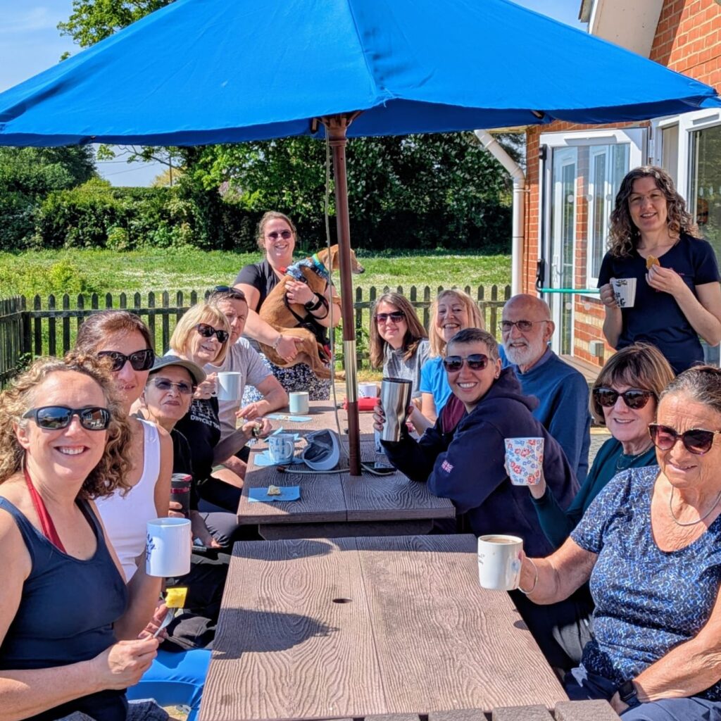 Members are enjoying cups of tea and pieces of cake after a paddle. They are sitting outside the club house on a long picnic table. There is a picnic umbrella up. Everyone is looking towards the camera and smiling.