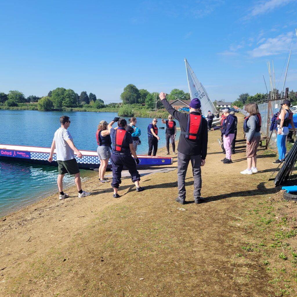 OPfL members are standing on the edge of the lake doing warm up exercises. The boat is in the water, the sky and the lake are blue, the sun is shining.