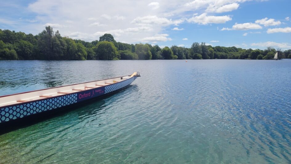 The dragonboat is empty and sitting on the edge of the lake. The lake is blue, there are clouds in the sky, and the sun is shining.