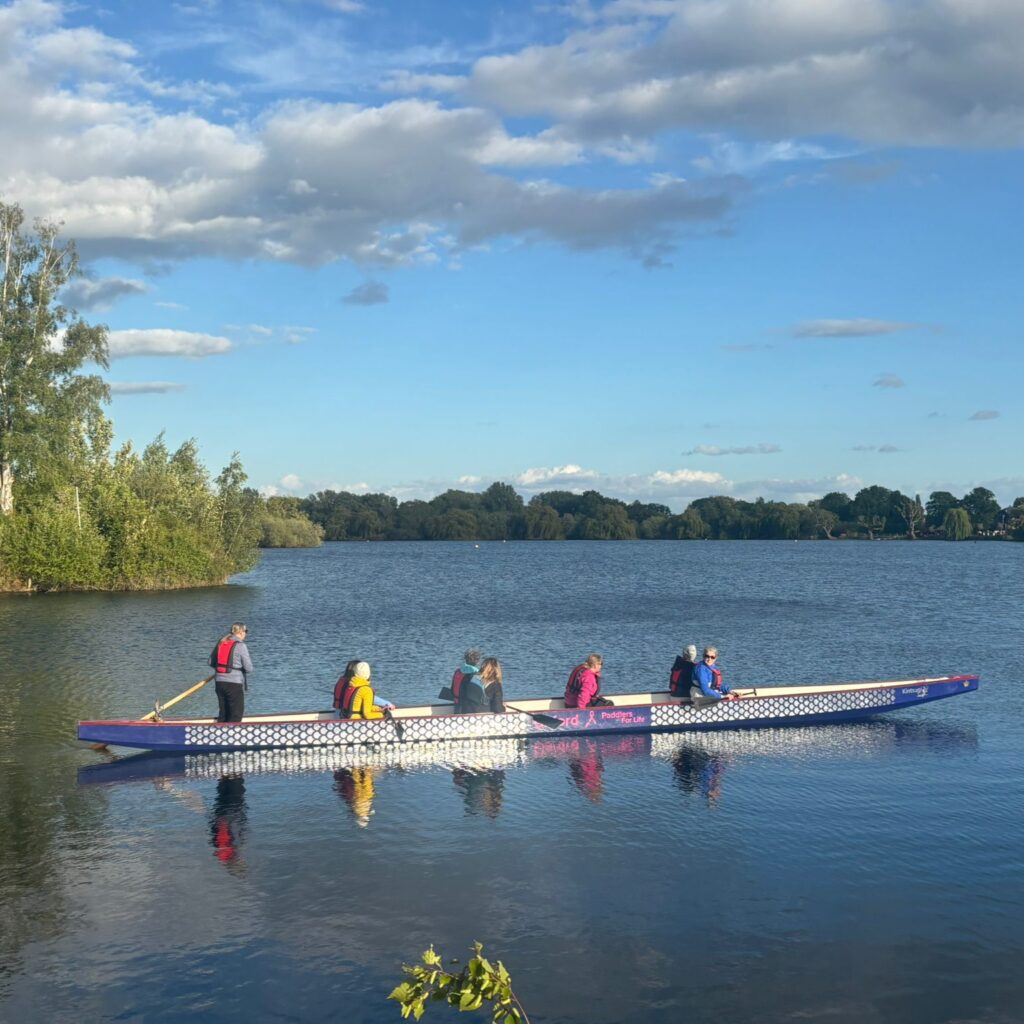 The dragonboat is out on the water. We can see the helm standing at the back. There are 7 people in the boat. Behind the boat is the tree line and the sky.