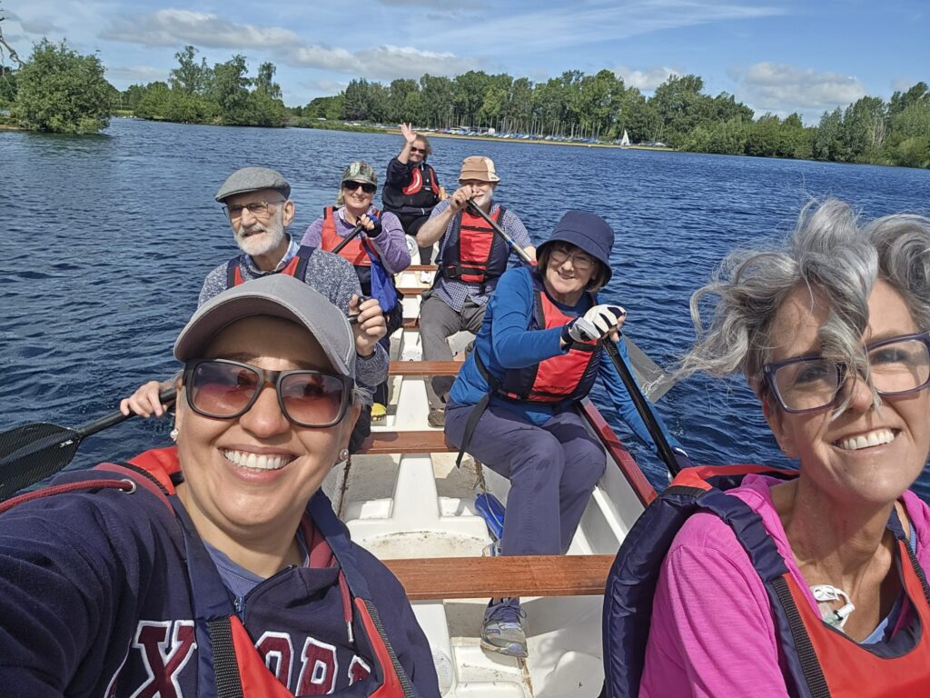 7 paddlers are in the boat. The helm is waving at the camera, people are smiling. In the background we can see the tree line at the edge of the lake, and the boats at the sailing blue.