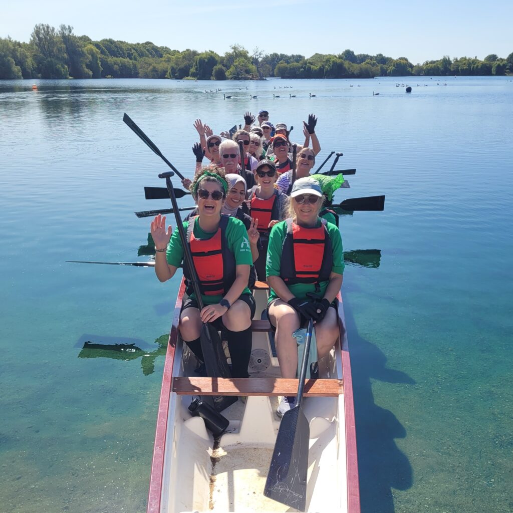 Paddlers are sitting in the boat on the lake waving at the camera. The weather is sunny and the sky is blue.